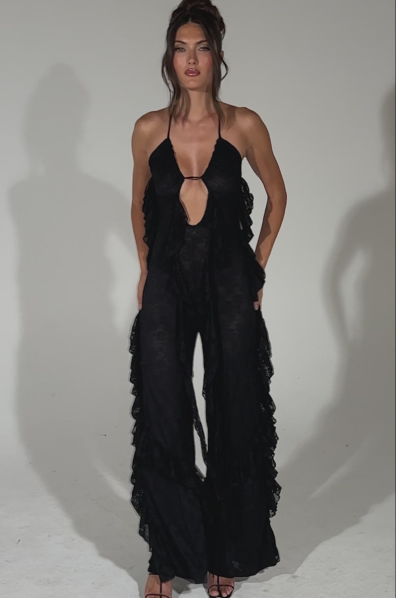 A video of a woman modeling the FREE AS A BIRD RUFFLED JUMPSUIT—a black halter jumpsuit with lace accents, ruffled details, a keyhole neckline, and wide-leg pants—paired with black high-heeled sandals against a light blue background.