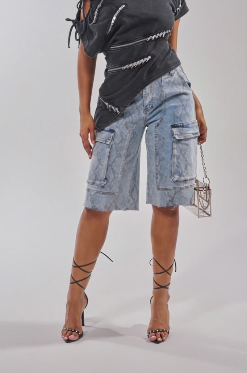 A video of a woman wearing the LITTLE SNAKE BERMUDA SHORT in light blue snake print, styled with large sunglasses, a black off-shoulder zippered top, strappy lace-up heels, and a small bag.