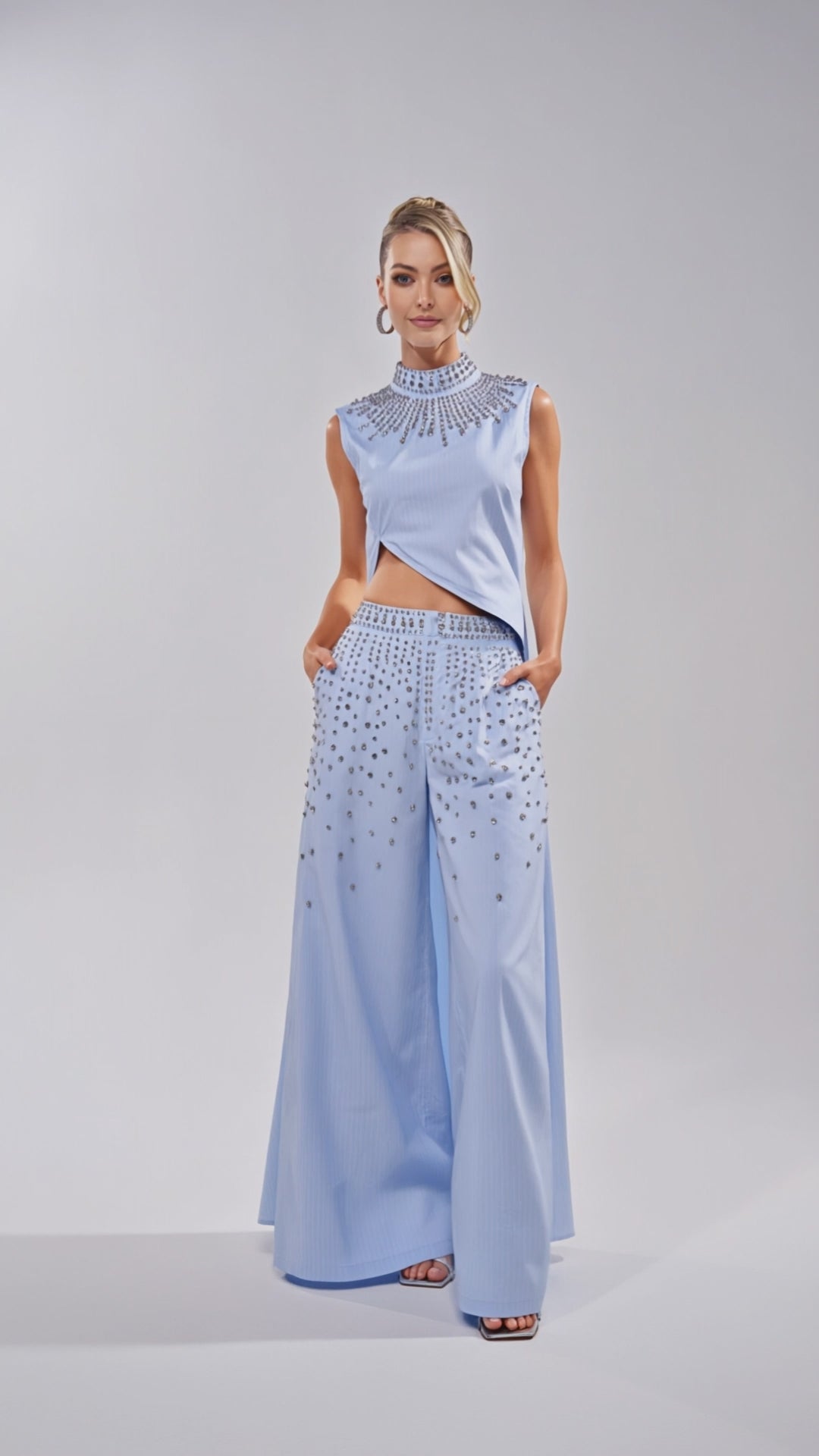Video of woman modeling the STAR OF THE NIGHT POPLIN BLOUSE, a sleeveless light blue crop top with a rhinestone collar and intricate beading, paired with matching wide-leg pants. Her hair is styled up against a plain, light background.