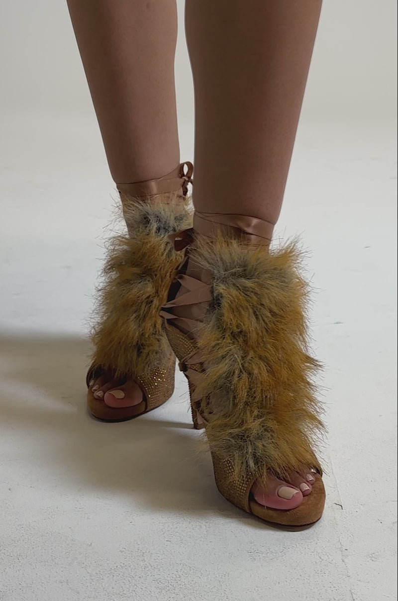 A video of a person wearing the AZALEA WANG MADHAV Bronze Rhinestone and Fox Fur Sandal, featuring thick brown and tan faux fur, an open toe, laced front, textured heels, and sparkling rhinestone details.