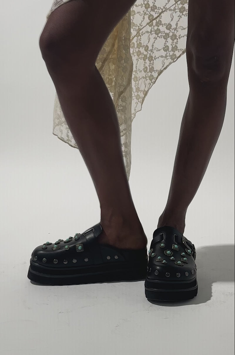 A video of a person wearing a cream lace dress and the AZALEA WANG MAVIS FLAT EMBELLISHED CLOG featuring turquoise stones, silver studs, and a western buckle.
