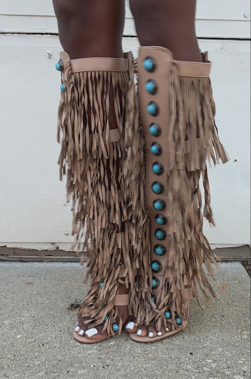 A video of a model wearing the AZALEA WANG AMADEO TAN SANDAL—tan, high-heeled, knee-high sandals with fringe and turquoise bead accents—this person stands on a concrete surface.
