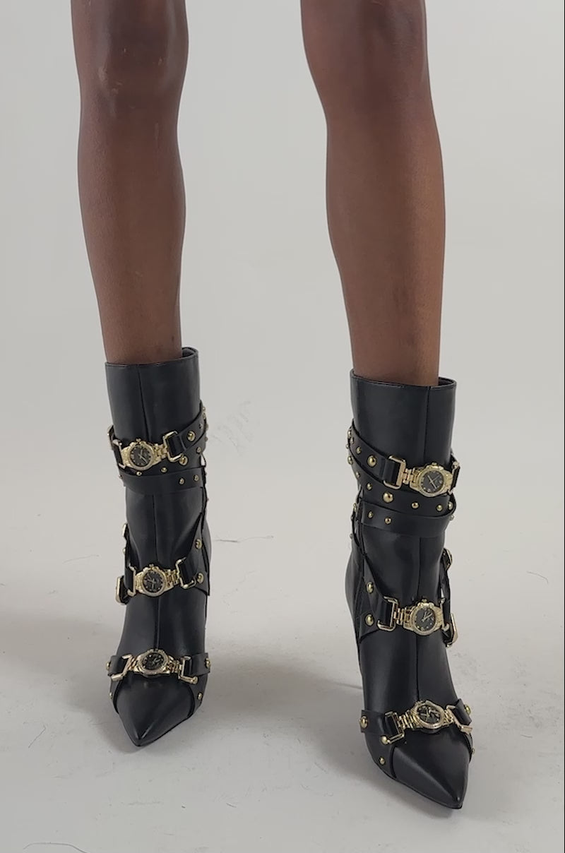 Video of a woman modeling the AZALEA WANG WATCH ME BOOTIE WITH WATCHES—black faux leather boots adorned with gold buckles and embellishments.