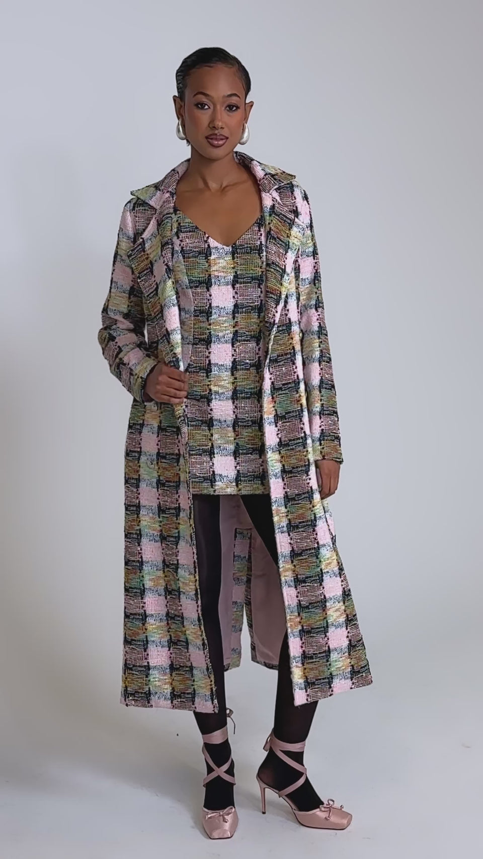 A video of a woman in the MAIN SQUEEZE TWEED MINI DRESS with a long, multicolored coat, black tights, and pink ballet flats against a plain, well-lit background.