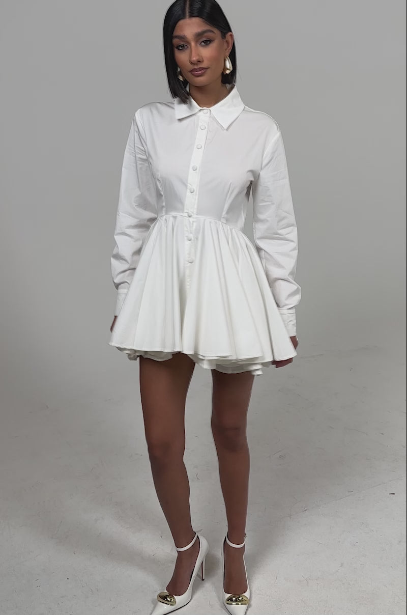 A video of a woman wearing THE IT GIRL POPLIN MINI DRESS—featuring long sleeves, a collared button-up top, flared pleated skirt—and pointed-toe white high heels.