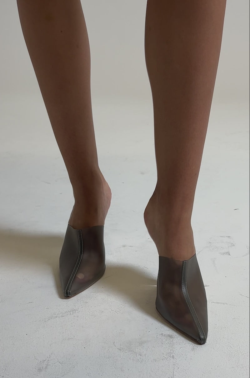 A video of a person wearing the AKIRA GOLD SANCTUS GREY PVC PUMP—clear pointed-toe heels with transparent wedge—paired with a pinstriped skirt, standing on a light-colored surface.