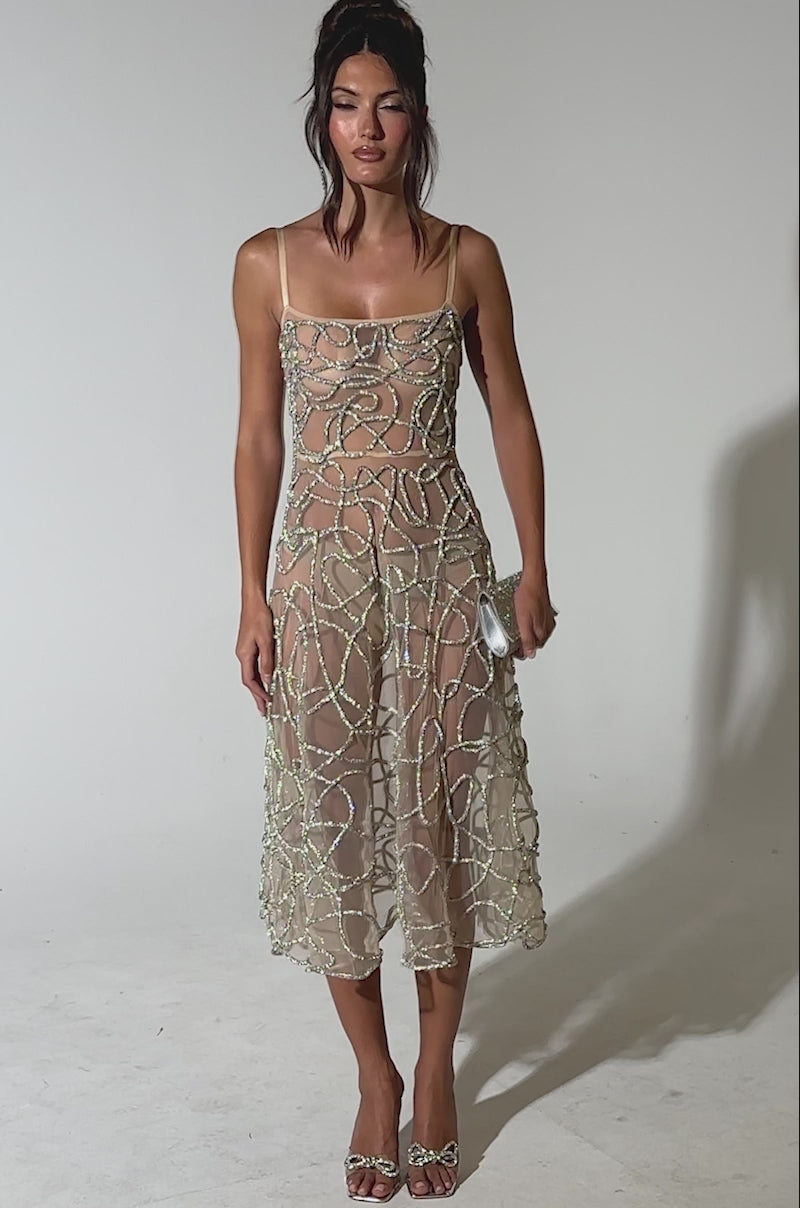 A video of a model wearing the RIGHT THIS WAY RHINESTONE MIDI DRESS IN NUDE, featuring sheer mesh and glittering bead details, styled with a silver clutch and silver open-toe heels against a light blue background.
