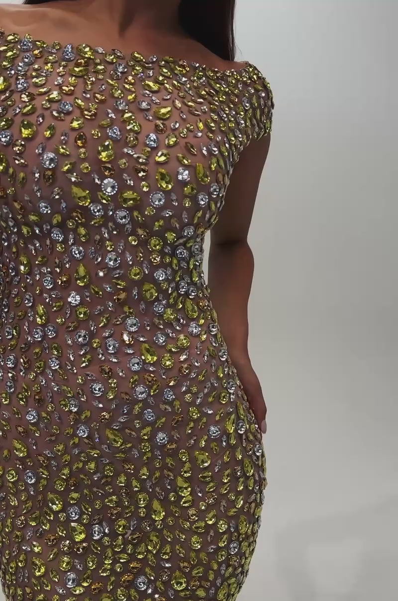 A video of a woman wearing the HIGH STANDARDS RHINESTONE MINI DRESS, featuring an off-shoulder, sheer nude bodycon silhouette enhanced with yellow and silver rhinestone embellishments, accessorized with a clutch, long straight hair, and star-shaped earrings.