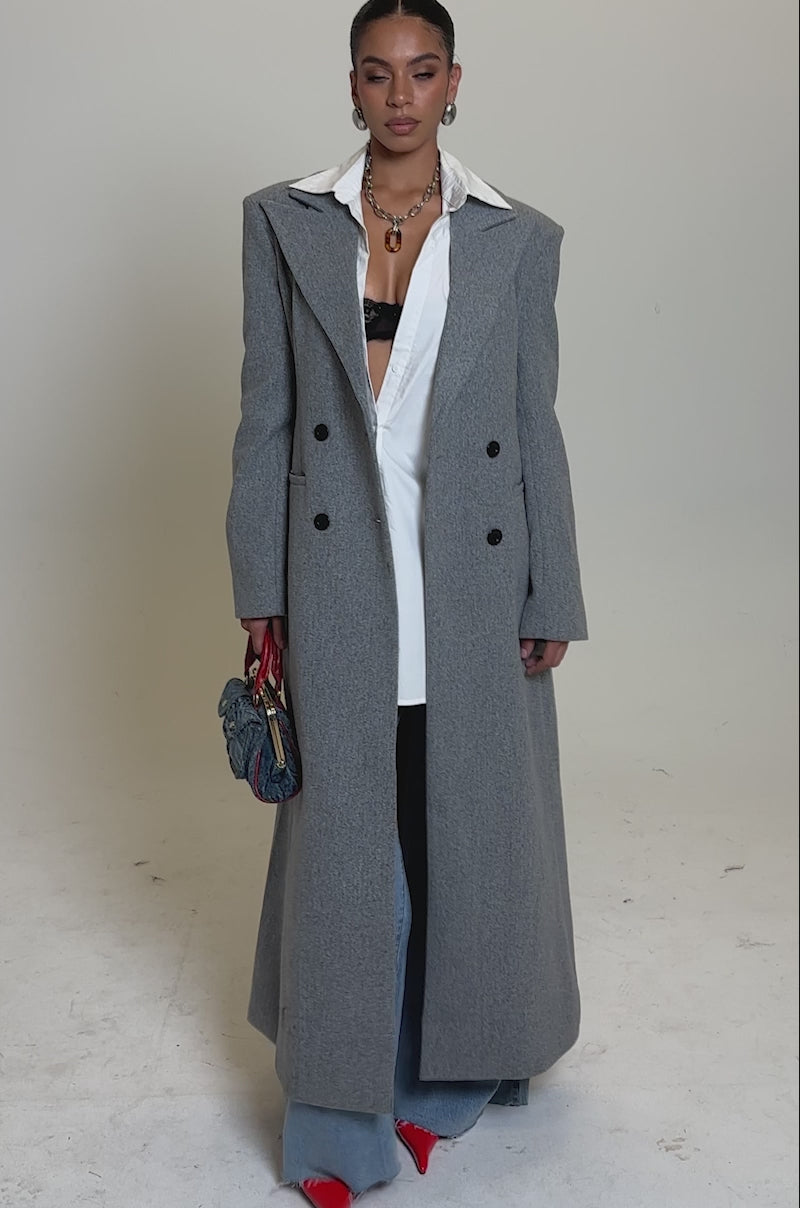 A video of a woman wearing the SCARLETT LUXE TRENCH COAT over a white shirt and blue jeans, holding a small denim handbag with red handles, paired with red pointed-toe shoes and statement earrings.