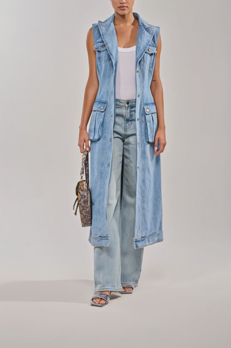 Video of woman modeling the GOTTA DIP LONG DENIM VEST, a sleeveless utility-inspired vest with front pockets, styled over wide-leg denim pants, open-toe heels, and a patterned handbag.