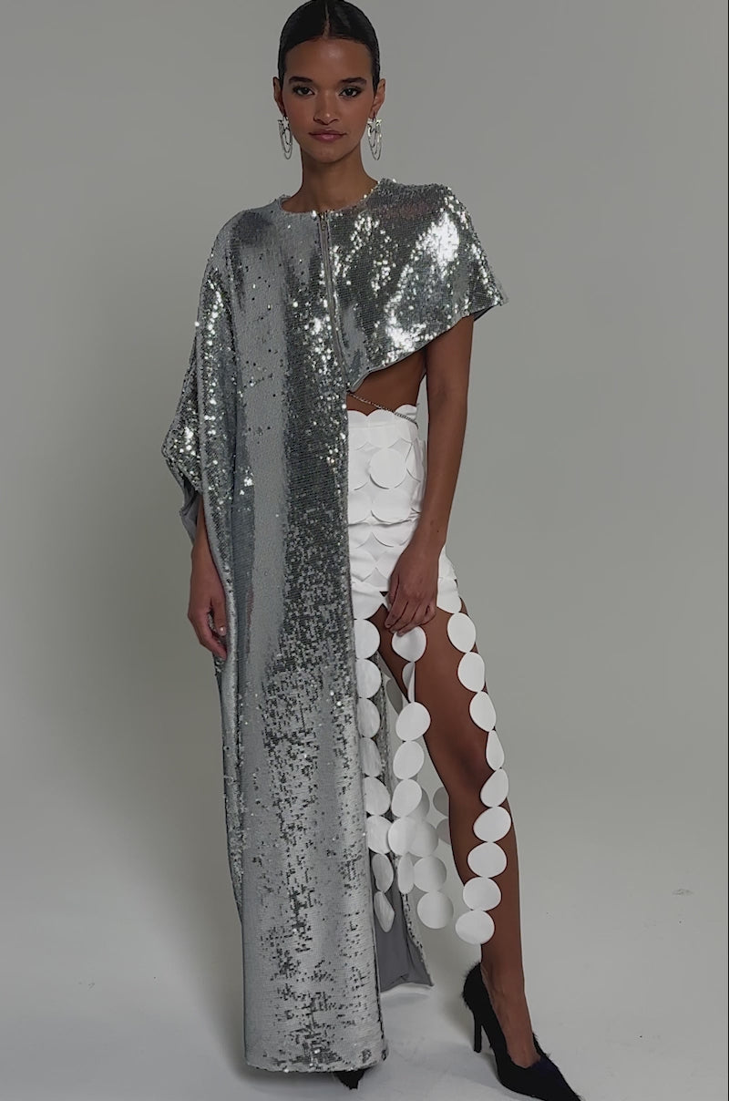 Video of woman modeling the MIDDLE NAME IS DRAMA HIGH LOW BLOUSE, a shiny silver metallic sequin cape-style top, over a white high-slit skirt. She pairs it with black heels and hoop earrings, hair pulled back, against a plain light background.