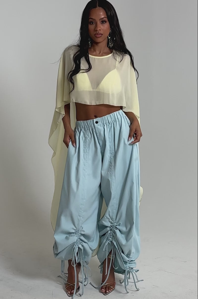 Video of woman modeling the DRAMA RUCHED JOGGER IN BLUE with adjustable ankle drawstrings, styled with sheer high-heeled sandals and a flowing yellow top—a chic, modern outfit ideal for resort vibes.