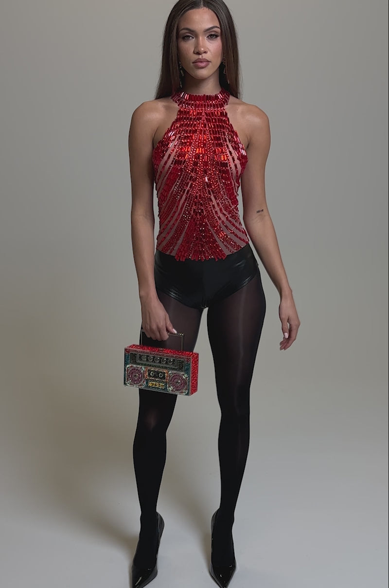 Video of woman modeling the FIRECRACKER RHINESTONE BLOUSE, a red sleeveless top with beads and sheer fabric, paired with high-waisted black pants. She holds a colorful clutch bag and gazes to the side