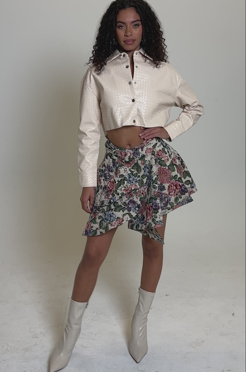 Video of woman modeling the GIVE ME FLOWERS MINI SKIRT—a high-waisted, vintage-inspired floral mini skirt featuring an asymmetrical ruffle layer and matching belt—against a plain, light background.
