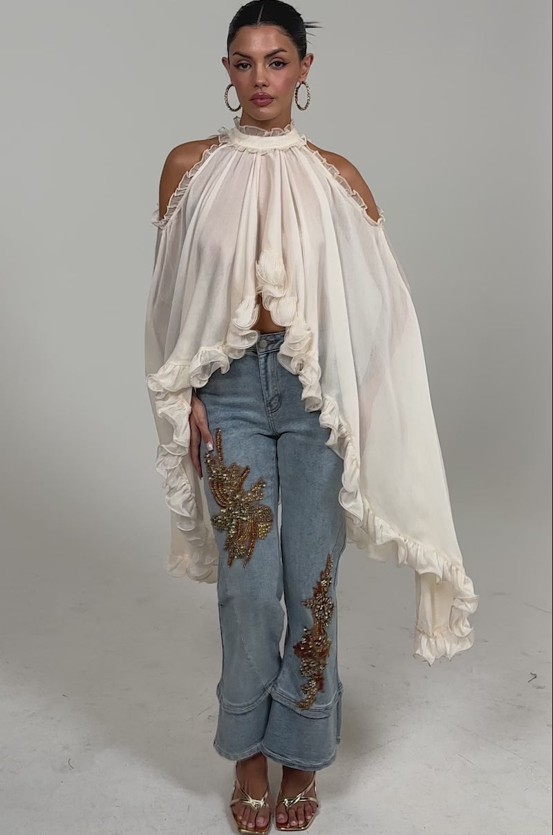 Video of woman modeling the TOTALLY COOL RELAXED DENIM featuring gold beaded embroidery on both legs, styled with gold sandals and a sheer, ruffled cream top.