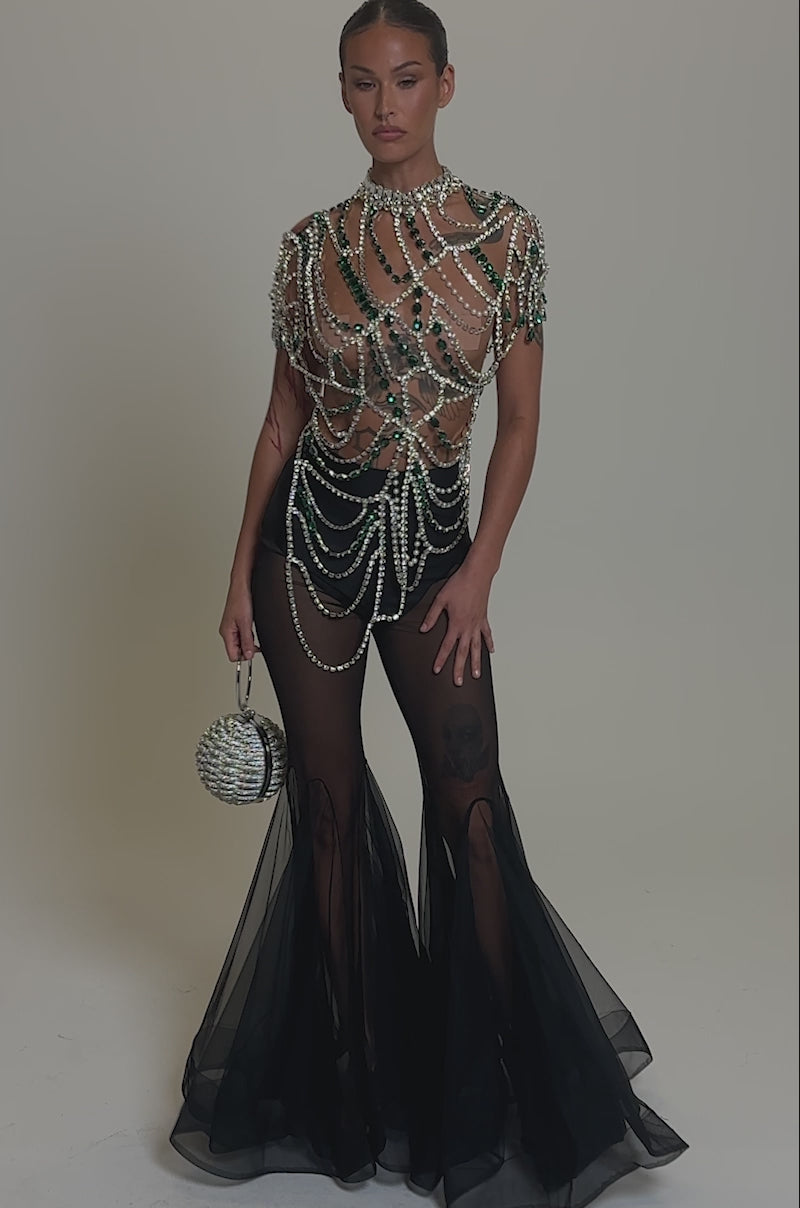 Video of woman modeling the DANCING QUEEN RHINESTONE CHAIN TOP with sparkling green and silver gems, black sheer pants, and a round silver beaded bag, she poses confidently with slicked-back hair and a serious expression for a bold, standout look.