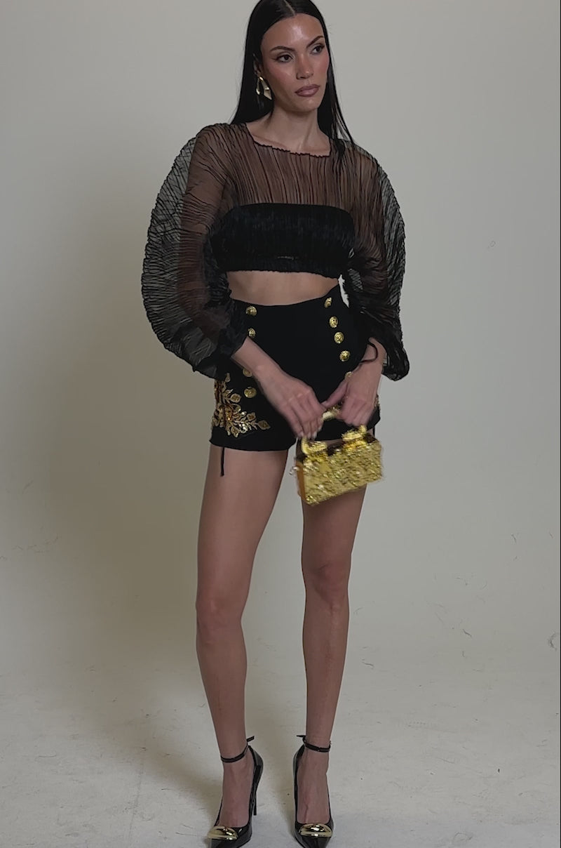 A video of a woman wearing the FORTUNES FAVORITE SHEER LONG SLEEVE CROP TOP with pleated details and voluminous sleeves, high-waisted black shorts with gold accents, black heels, and holding a chic small gold handbag.