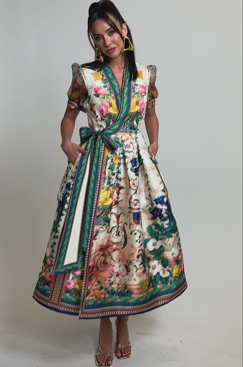A video of a woman in the FLOWER GIRL SATIN MAXI DRESS, a botanical print wrap maxi with puff sleeves and a waist sash. 