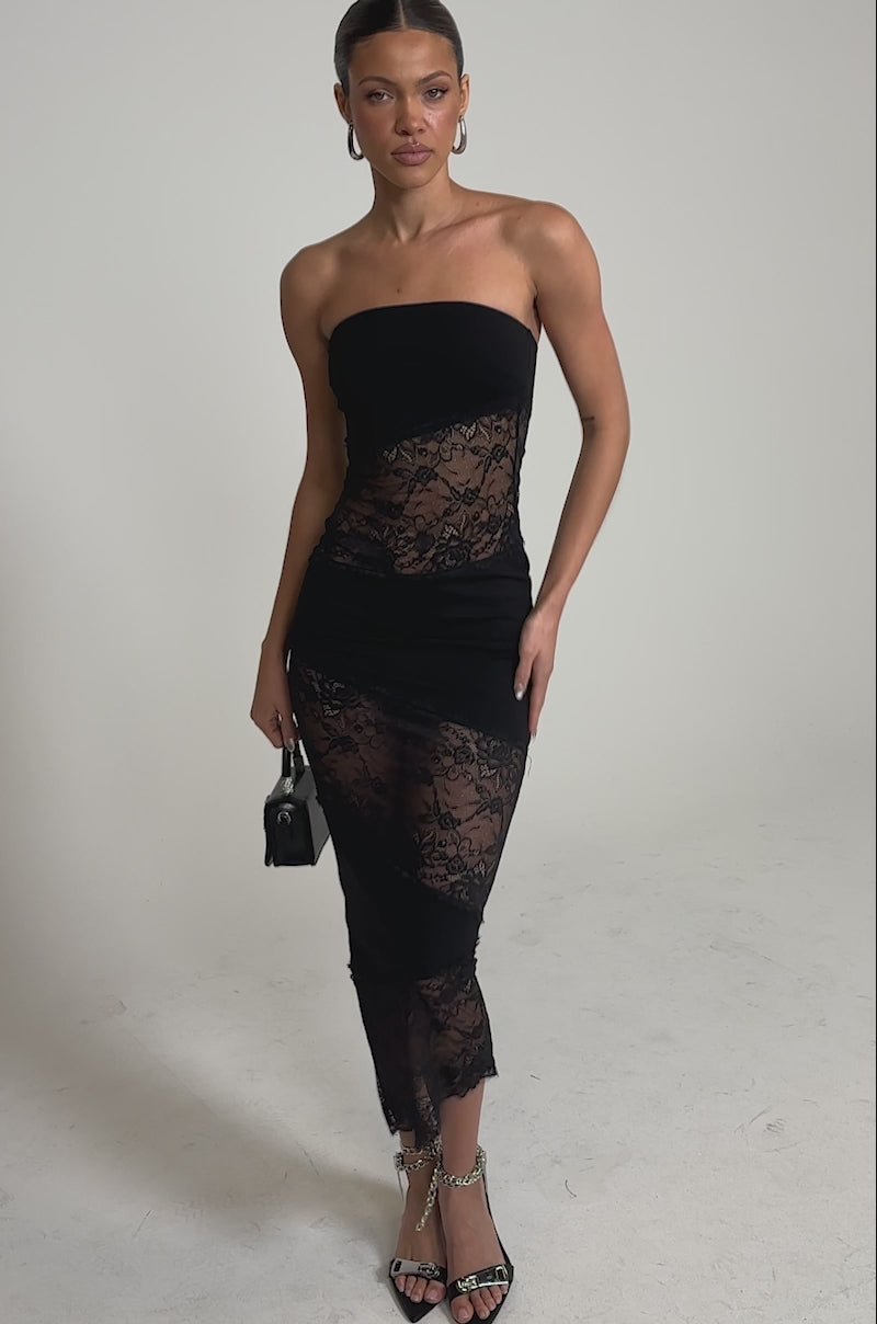 A video of a woman in the ILL SHOW YOU LACE MAXI DRESS, featuring strapless lace, sheer panels, and an asymmetrical hem, holds a black handbag and wearing high-heeled sandals with silver chain details against a plain light background.