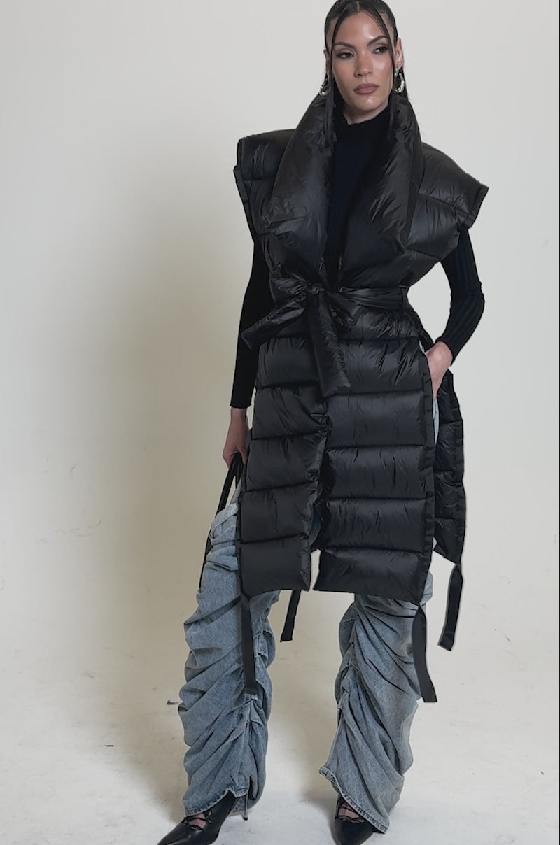 Video of woman modeling the PILOT JONES LONG LINE PUFFER VEST IN BLACK with a hood, layered over a black turtleneck, blue ruched jeans, and pointed shoes, carrying a black handbag against a plain gray background.