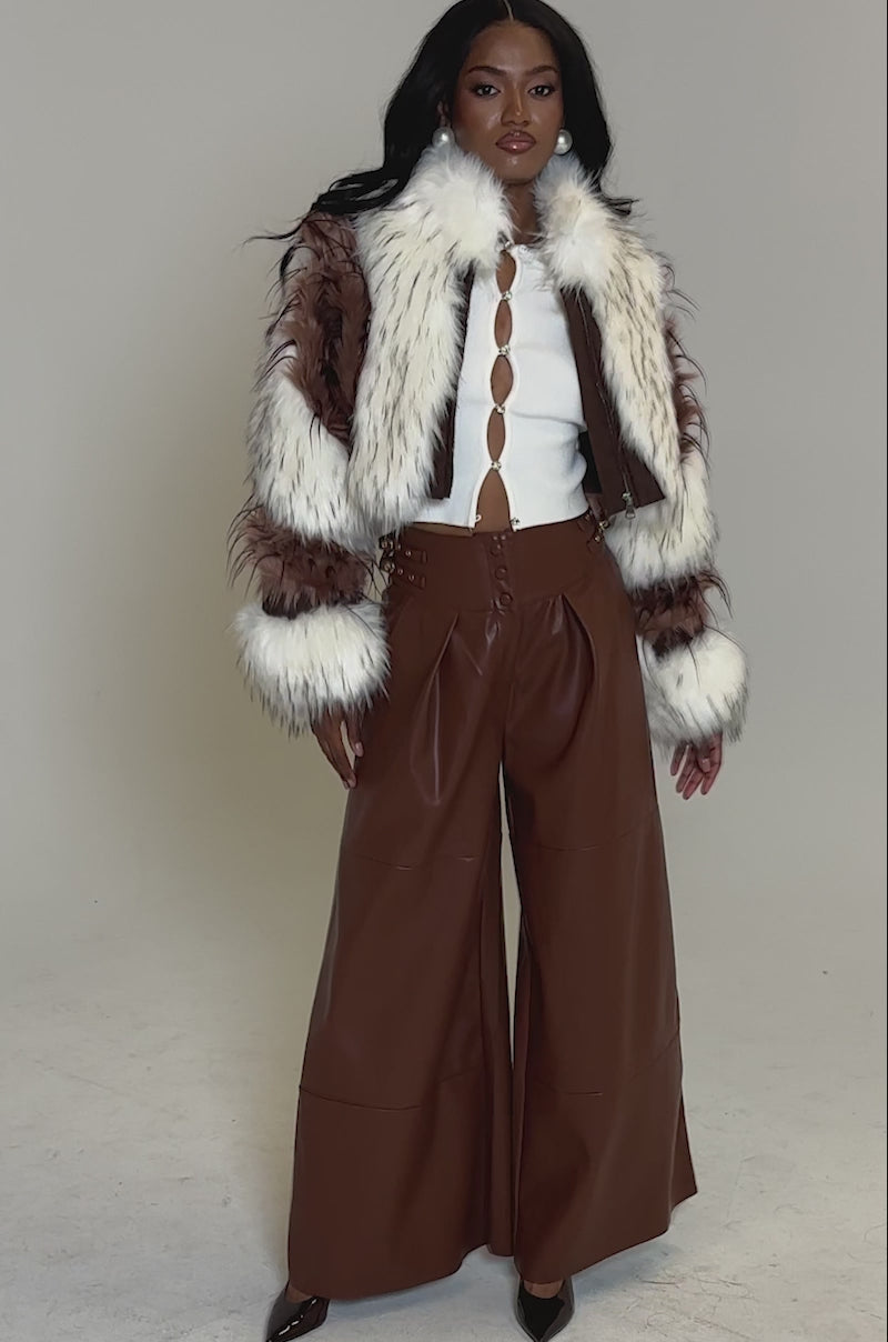 A video of a woman posing in the FRANKIE PATCHWORK FAUX FUR jacket, wide-legged brown leather pants, a white cut-out top, and brown heels against a plain gray background.