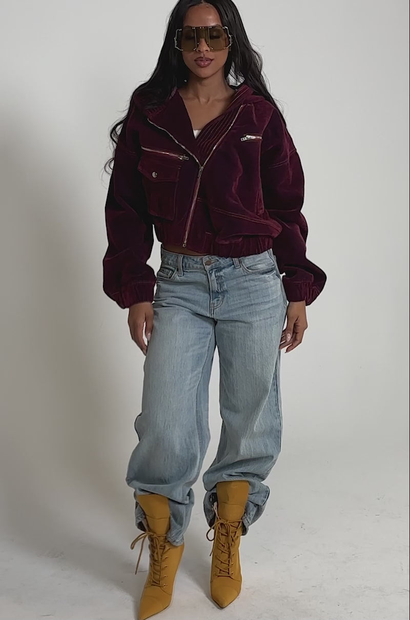 Video of woman modeling the PAYTON VELVET DENIM BOMBER—a maroon velvet jacket with an oversized hood—paired with large sunglasses and light blue jeans