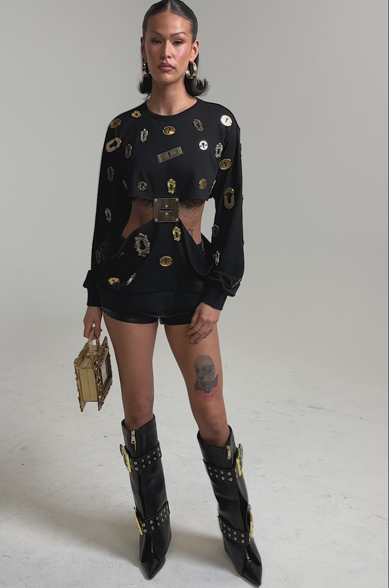 A video of a woman in bold streetwear: the KEY CHAIN TOP with gold keyhole motifs and metallic accents, paired with black shorts, tall boots with gold buckles, a gold box handbag, sleek makeup, and hoop earrings.