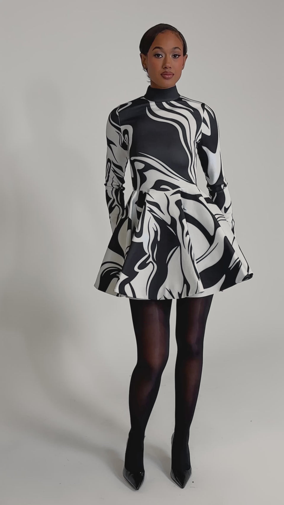 A video of a person wearing the GROOVY BABY MOCKNECK MINI DRESS, showcasing its black and white abstract design, styled with black sheer tights and high heels, set against a plain light background.