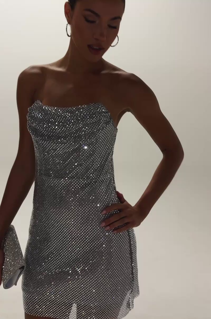 Video of woman modeling the ELECTRIC AURA RHINESTONE STRAPLESS MINI DRESS IN SILVER with hoop earrings and a silver clutch standing against a plain light background.