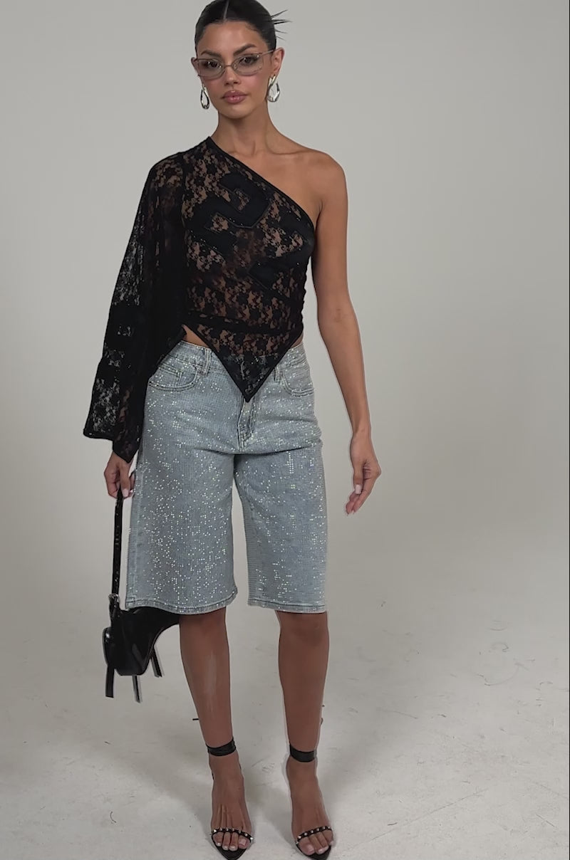 Video of woman modeling the GET IT DONE LACE JERSEY—a sheer black one-shoulder top with geometric details—paired with light blue jeans, hoop earrings, clear cat-eye glasses, and a sleek bun for a chic finish.