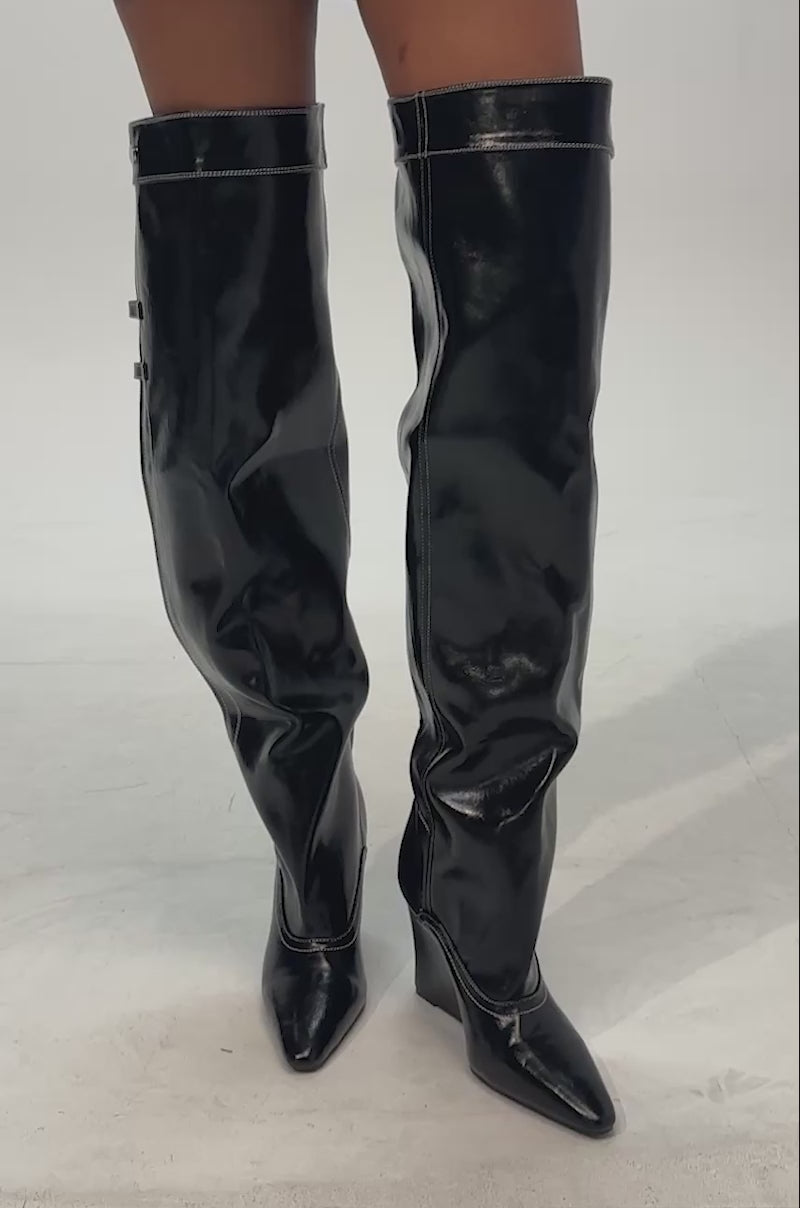 Video of woman modeling the AZALEA WANG RILO BLACK PU OVER THE KNEE WEDGE BOOT—a striking pair of shiny black, faux leather over-the-knee boots featuring pointed toes and visible stitching