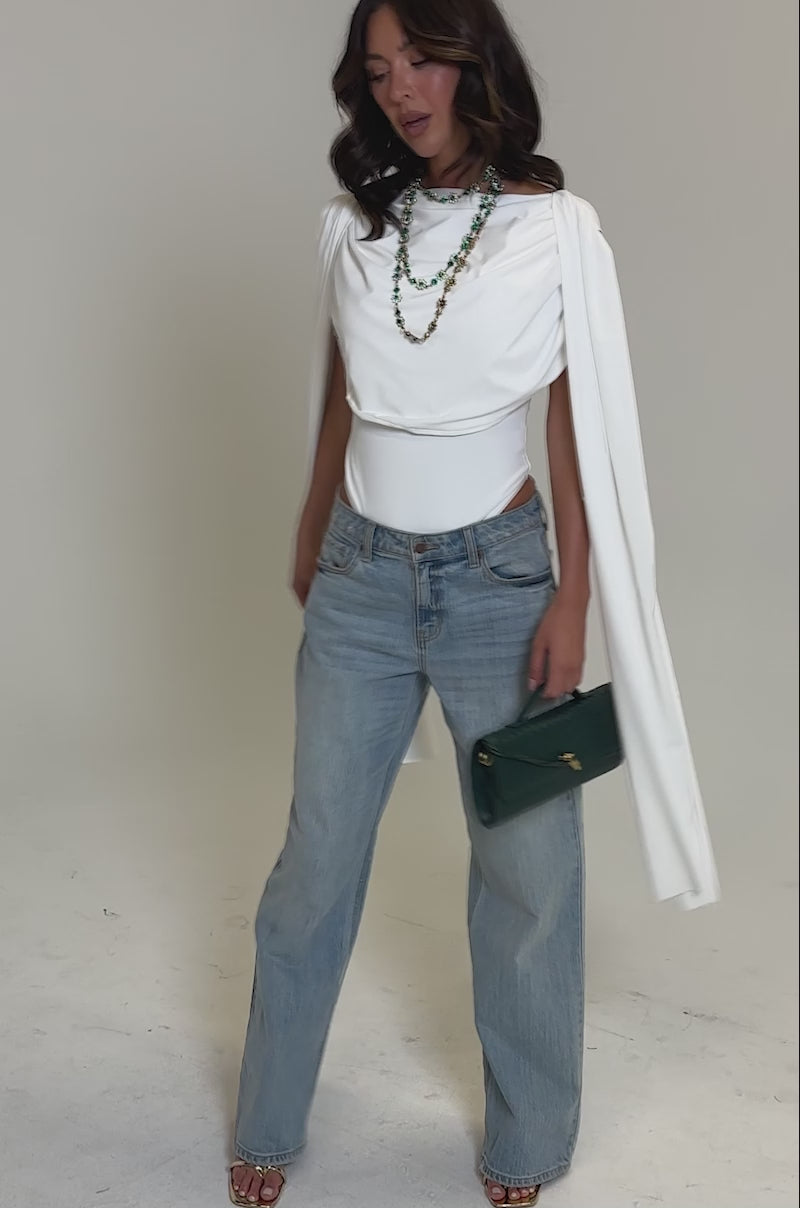 Video of woman modeling the HILLARY SEAMLESS BODYSUIT IN IVORY, light blue jeans, layered green bead necklaces, and holding a green quilted handbag.
