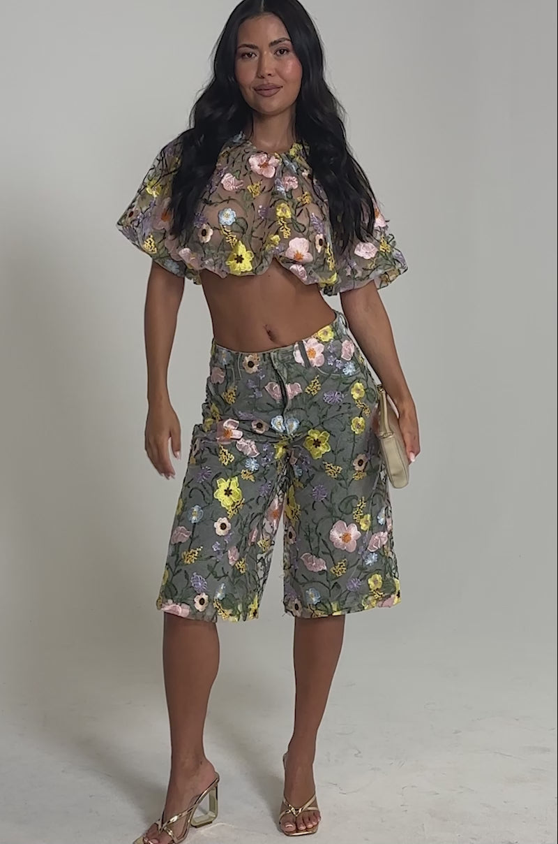 Video of woman modeling the GARDEN PARTY Stretch Denim with Floral Tulle Bermuda Shorts, paired with a sheer ruffled crop top. She accessorizes with a small clutch and heeled sandals against a plain, light-colored background.