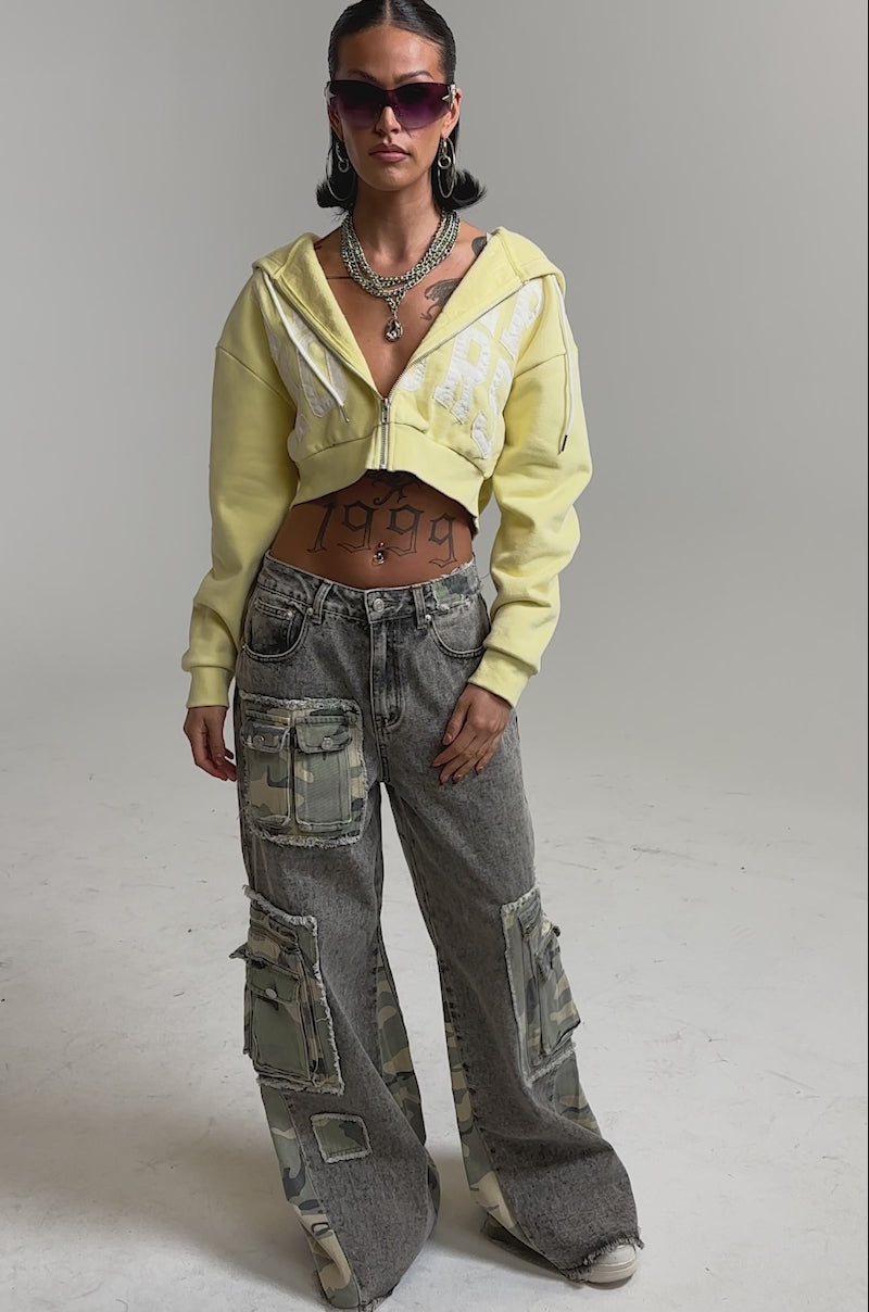 A video of a woman in the DOWN AND DIRTY CAMO CARGO jeans, yellow cropped hoodie, white sneakers, layered necklaces, and purple sunglasses.