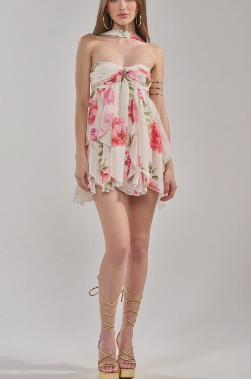 Video of woman modeling the TALK LATER FLORAL CHIFFON MINI DRESS, a strapless mini with pink rose and green leaf print, layered ruffles, matching neck scarf, gold arm cuffs, and long hair—perfect for a spring date night.