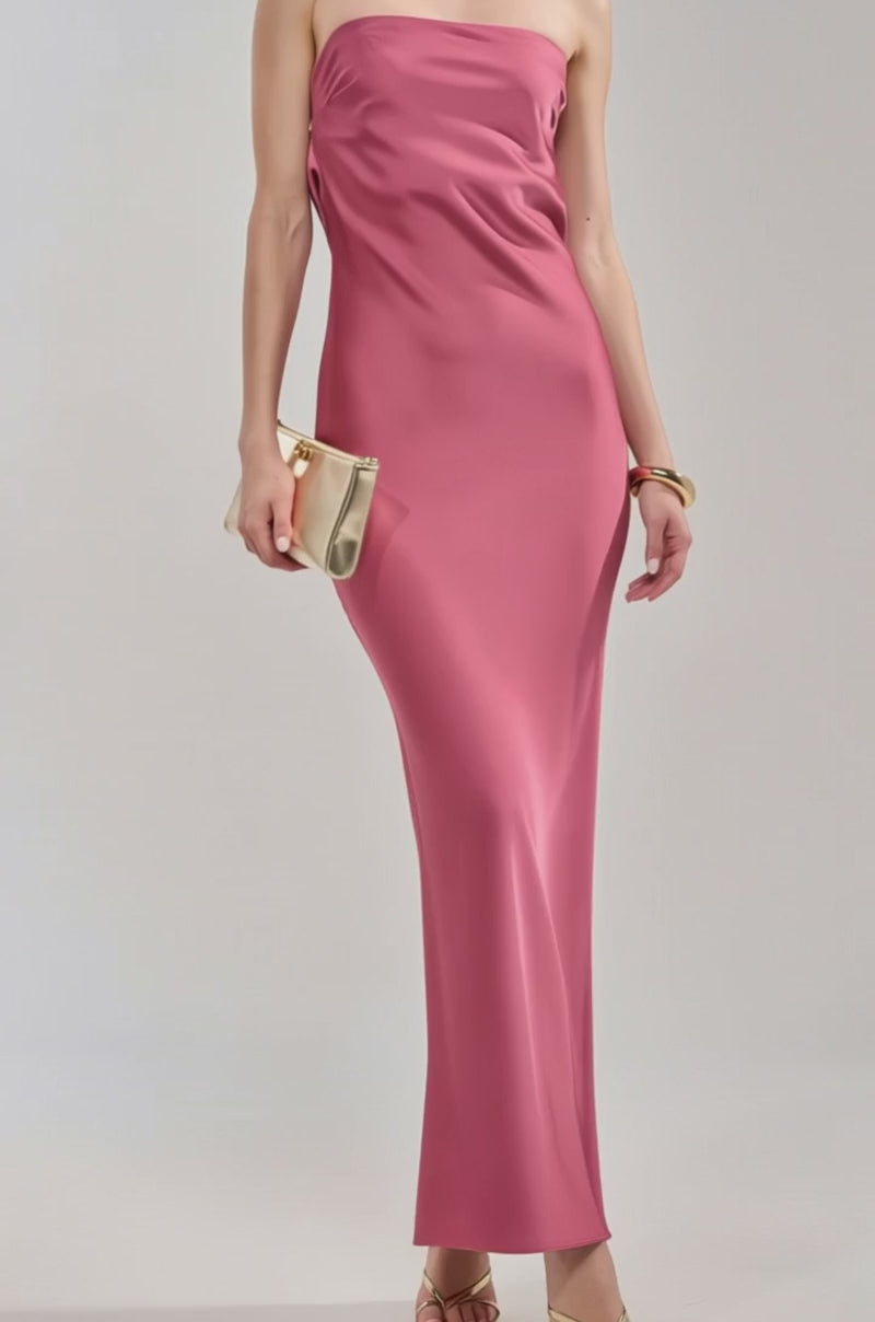 Video of woman modeling the SATIN SINNER MAXI DRESS, a rose pink satin gown with a draped back—ideal for weddings—holding a gold clutch. She accessorizes with gold heels 