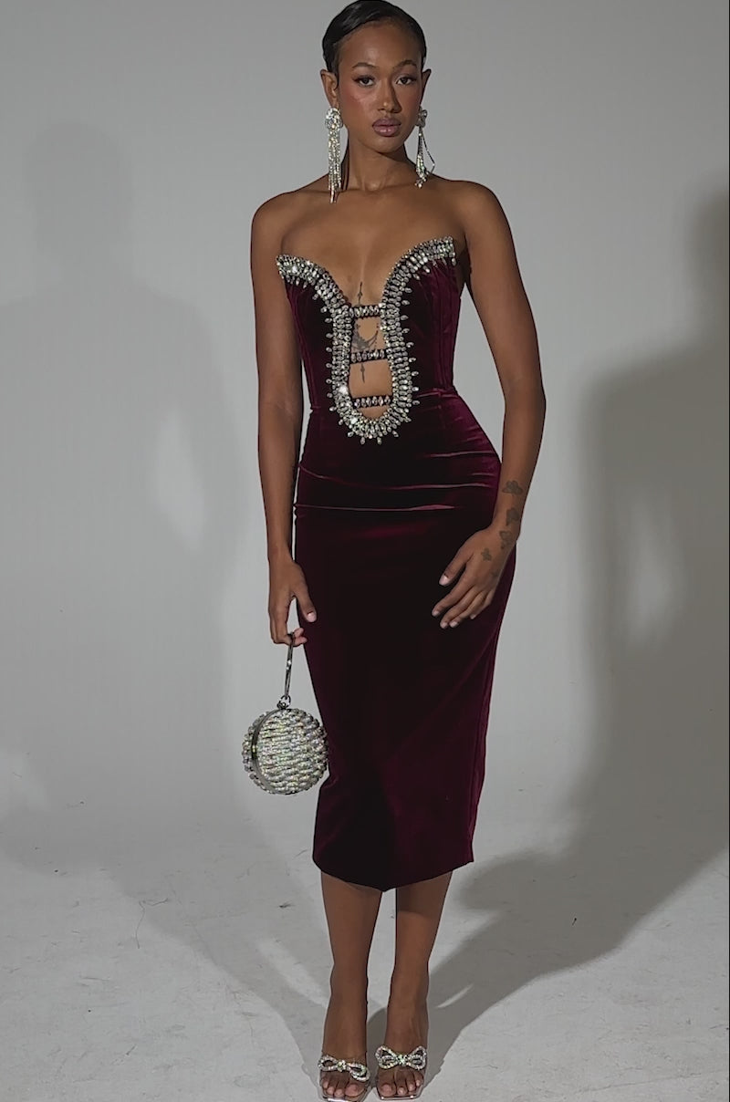 A video of a woman modeling the FEELIN THE LOVE VELVET RHINESTONE MINI DRESS, a strapless deep red velvet bodycon with bold ladder cutouts and sparkling rhinestones. She pairs it with a round beaded handbag, silver heels, and long earrings against a plain white background.