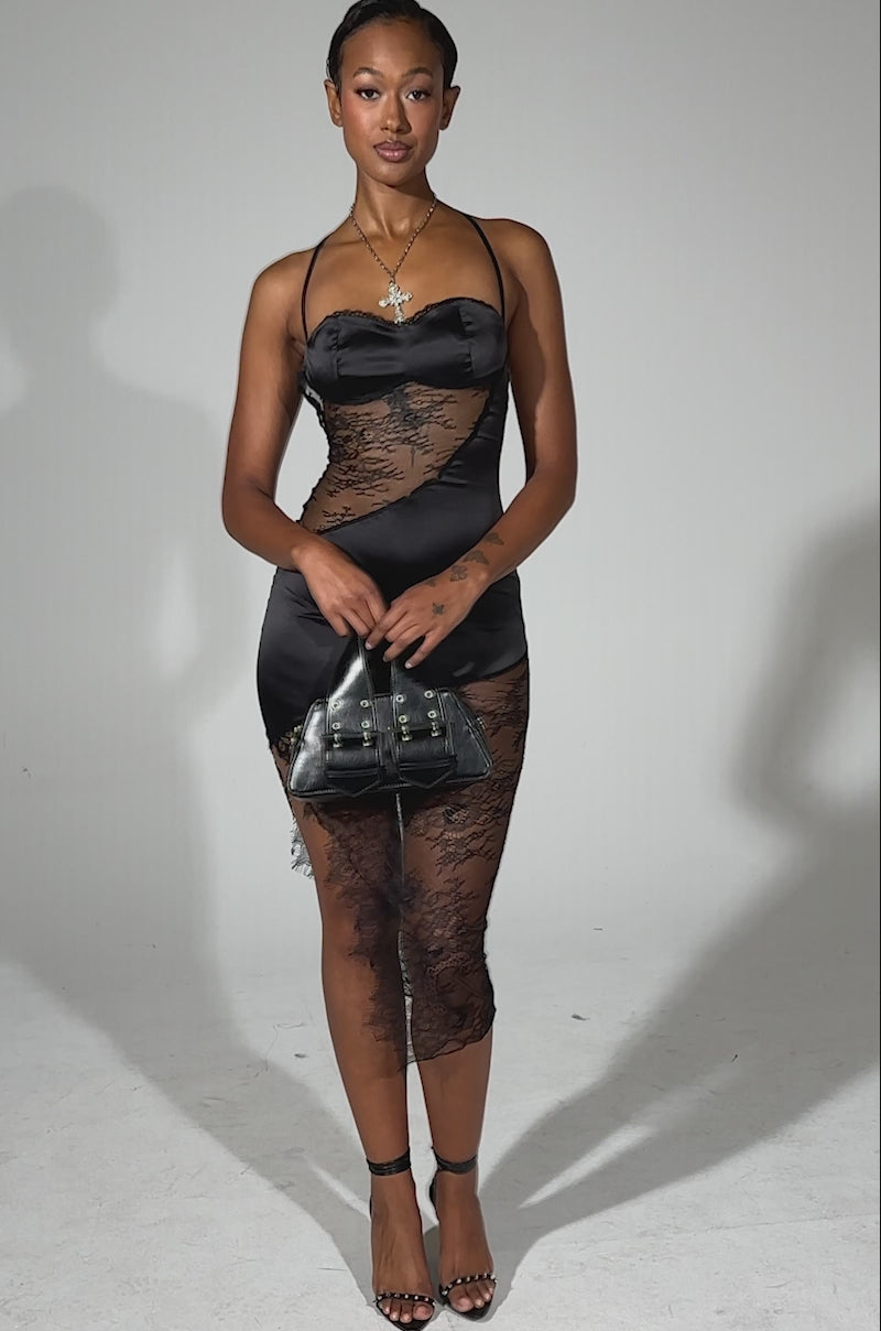 A video of a woman posing in the FEELIN MYSELF LACE CUTOUT MIDI DRESS, featuring black satin, lace details, sheer panels, and an asymmetrical design. She carries a black handbag and wears heels and a necklace with her hair slicked back.
