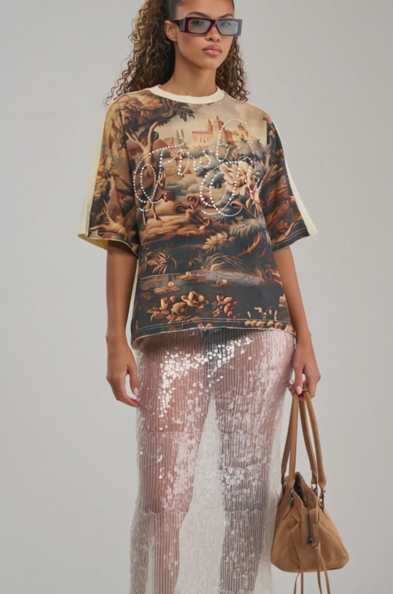 A video of a woman wearing sunglasses, the GOOD DAYS RHINESTONE GRAPHIC TEE featuring a detailed landscape print and rhinestone lettering, and a shiny, textured skirt holds a brown bag while standing against a plain background.