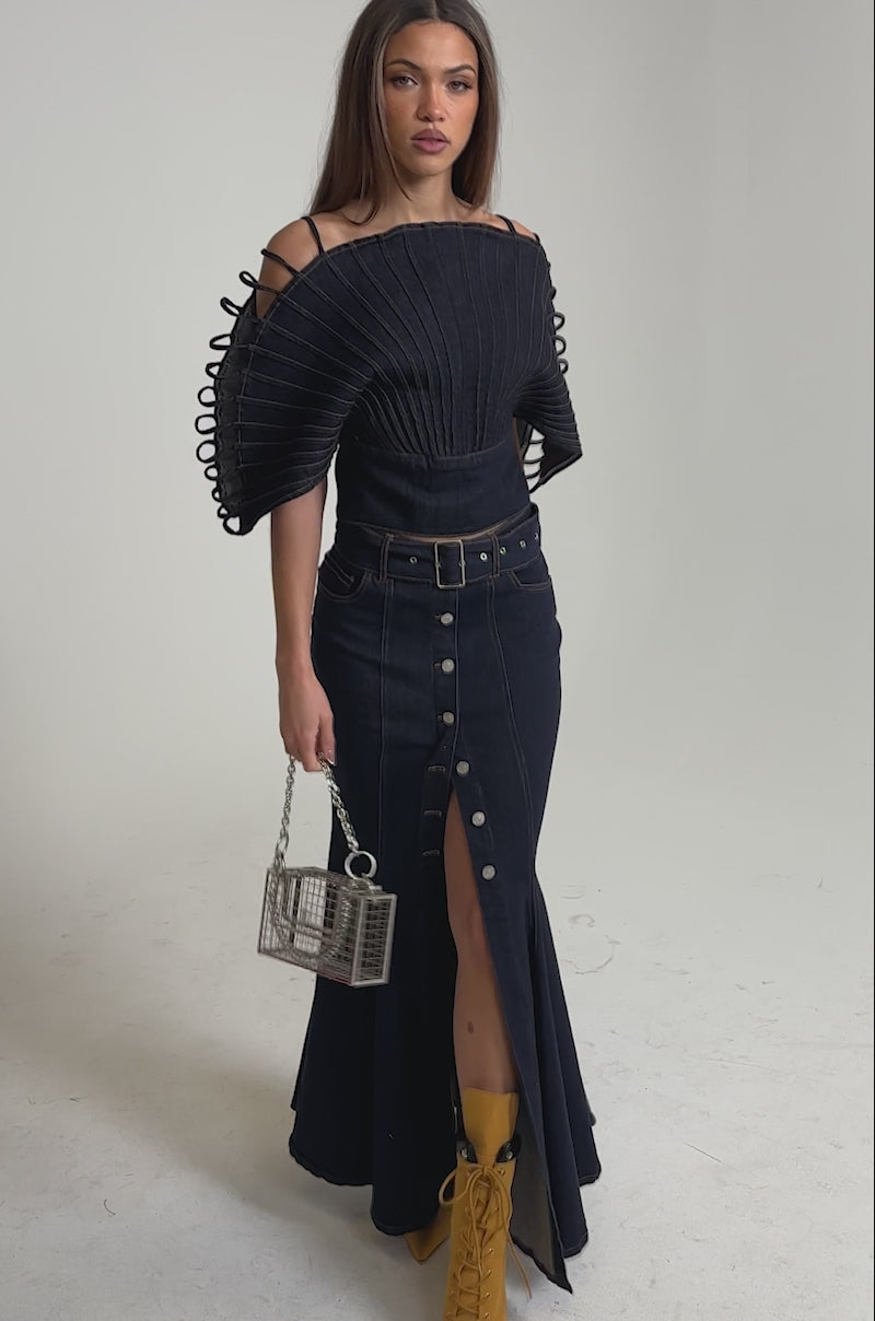 Video of woman modeling the DENIM ON DENIM BLOUSE IN DARK BLUE DENIM with sculptural sleeves and looped details, paired with a button-up skirt and carrying a transparent box-shaped handbag with a chain strap.