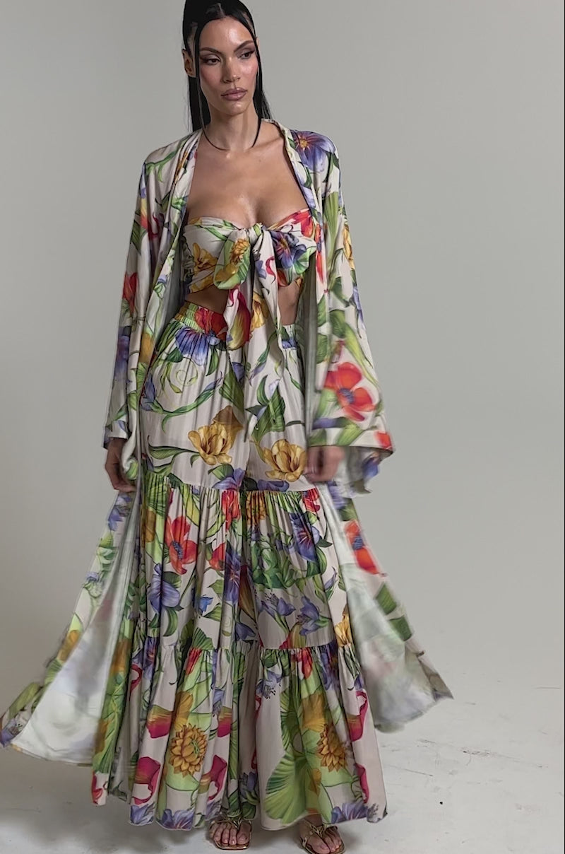 A video of a woman wearing the INTO THE GARDEN CARDIGAN in a longline silhouette with a matching tropical floral bandeau and flowing skirt, styled with gold sandals against a plain, light background.