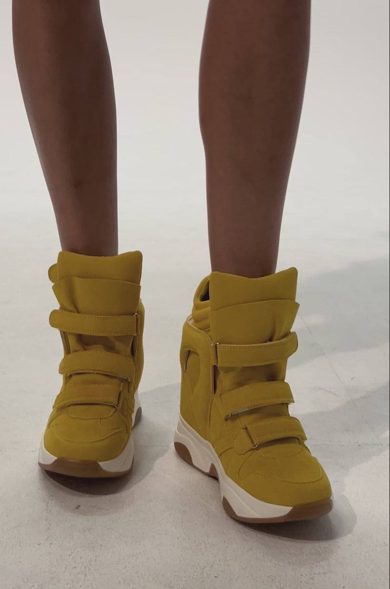 Video of woman modeling the AZALEA WANG MARANTO MUSTARD WEDGE SNEAKER—mustard yellow suede high-top sneakers with thick soles, velcro straps, and a hidden wedge heel against a plain light background.