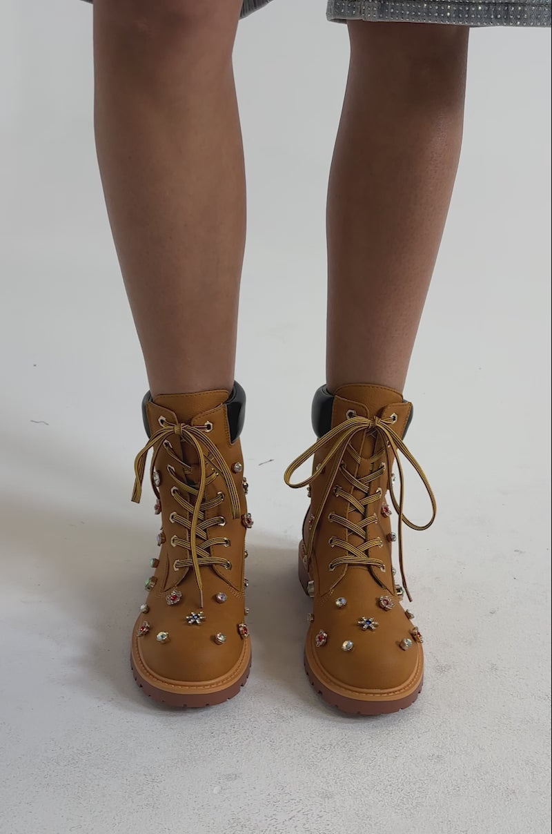 A video of a person wearing the AKIRA GOLD LUSTER TAN EMBELLISHED FLAT COMBAT FESTIVAL BOOTIE. 