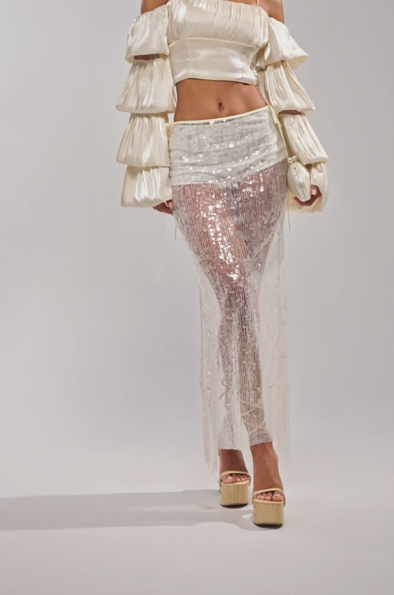 A video of a model wearing a cropped cream blouse with voluminous sleeves, the DRIPPING IN RHINESTONES MAXI SKIRT over matching bottoms, and beige platform sandals.