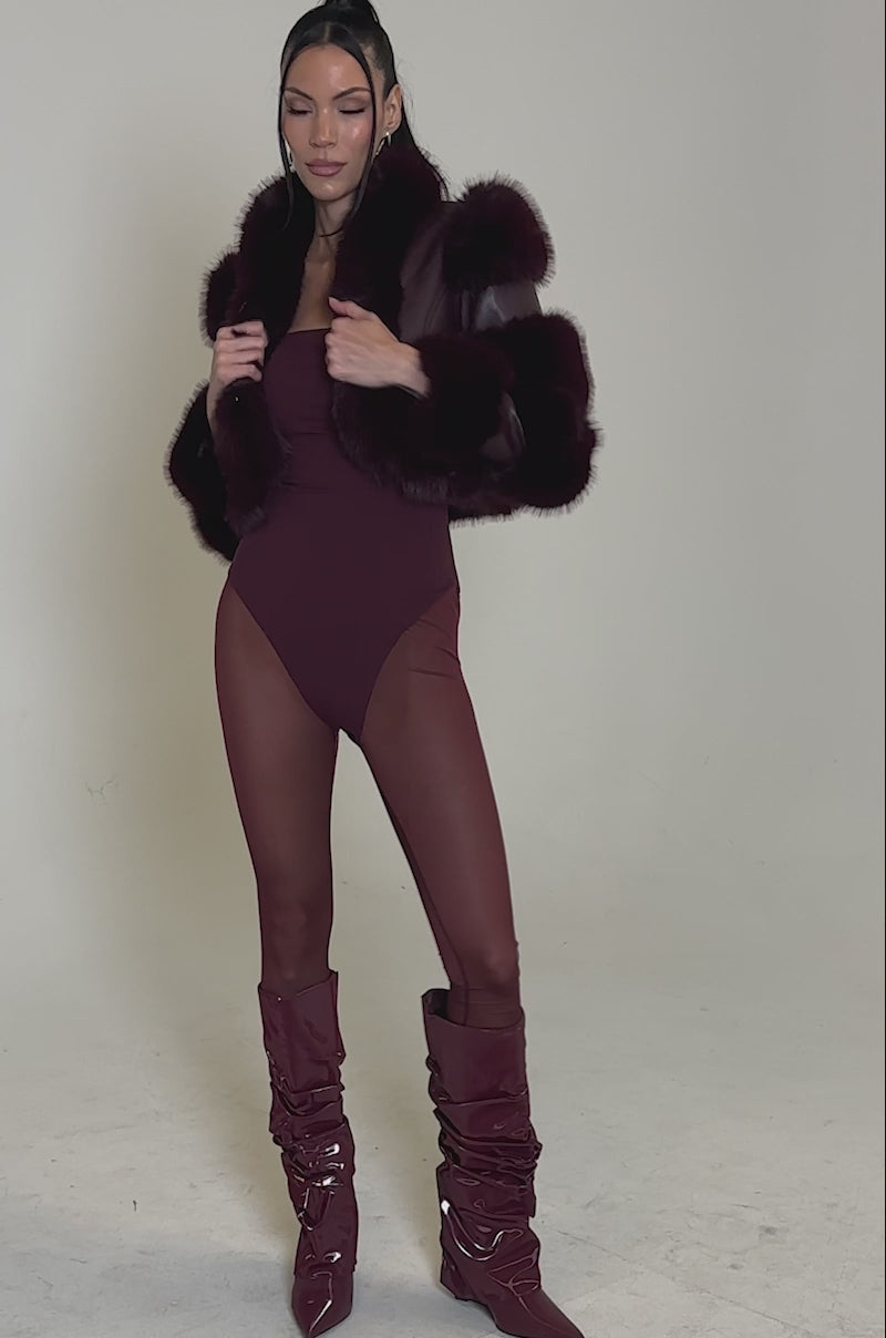 A video of a woman posing in the LOVERBOY FAUX FUR MOTO JACKET IN BURGUNDY, styled with a matching bodysuit, sheer tights, and slouchy leather boots. Her straight hair is tucked behind her ears against a plain light background.