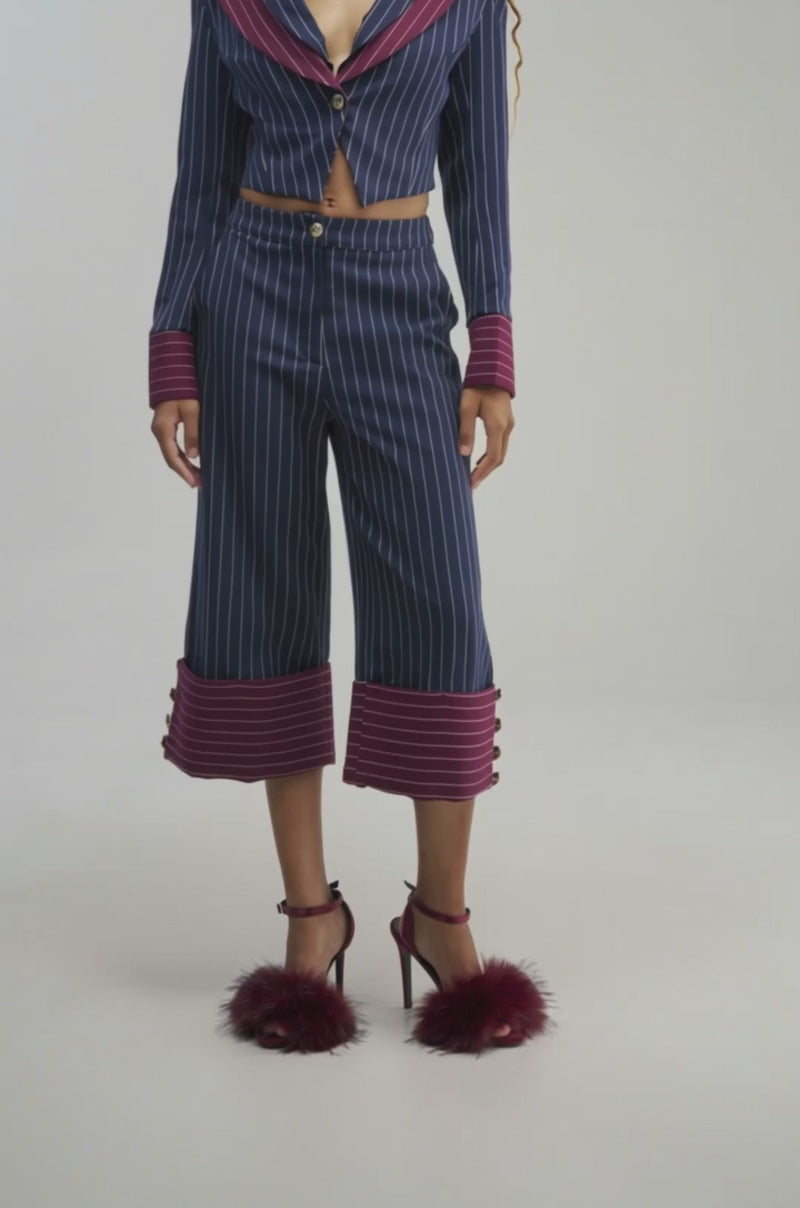 Video of a woman modeling the NEVER BETTER PINSTRIPE TROUSER featuring navy pinstripes and bold maroon cuffs, paired with a coordinating blazer detail and maroon high heels with plush accents. 