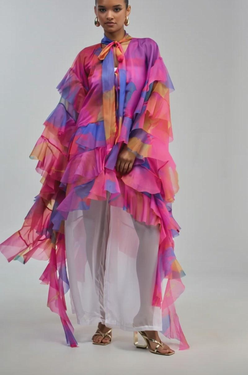 Video of a woman modeling the ELECTRIC SUNSET RUFFLE TOP, a flowing, sheer chiffon blouse featuring ruffled details and vibrant shades of pink, purple, and orange. 