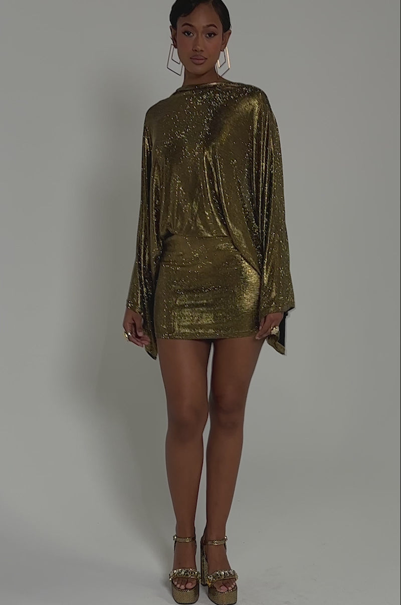 Video of woman modeling the GOLDEN HOUR RHINESTONE MINI DRESS, featuring a cape overlay and draped sleeves, paired with black heels against a dark background.
