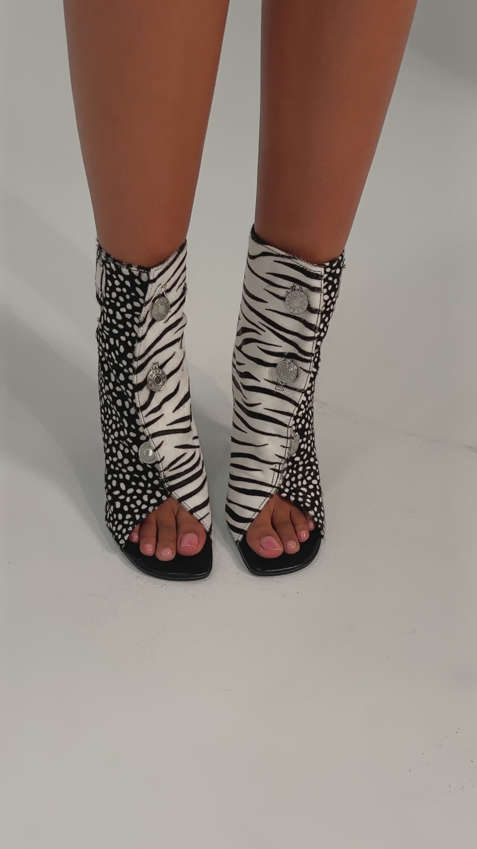 A video of a person wearing the AZALEA WANG DOROTHY BLACK CHEETAH CALF HAIR PRINTED STILETTO BOOTIE—featuring high heels, open toes, a black-and-white animal print, button details, and a spotted side—against a plain light-colored background.