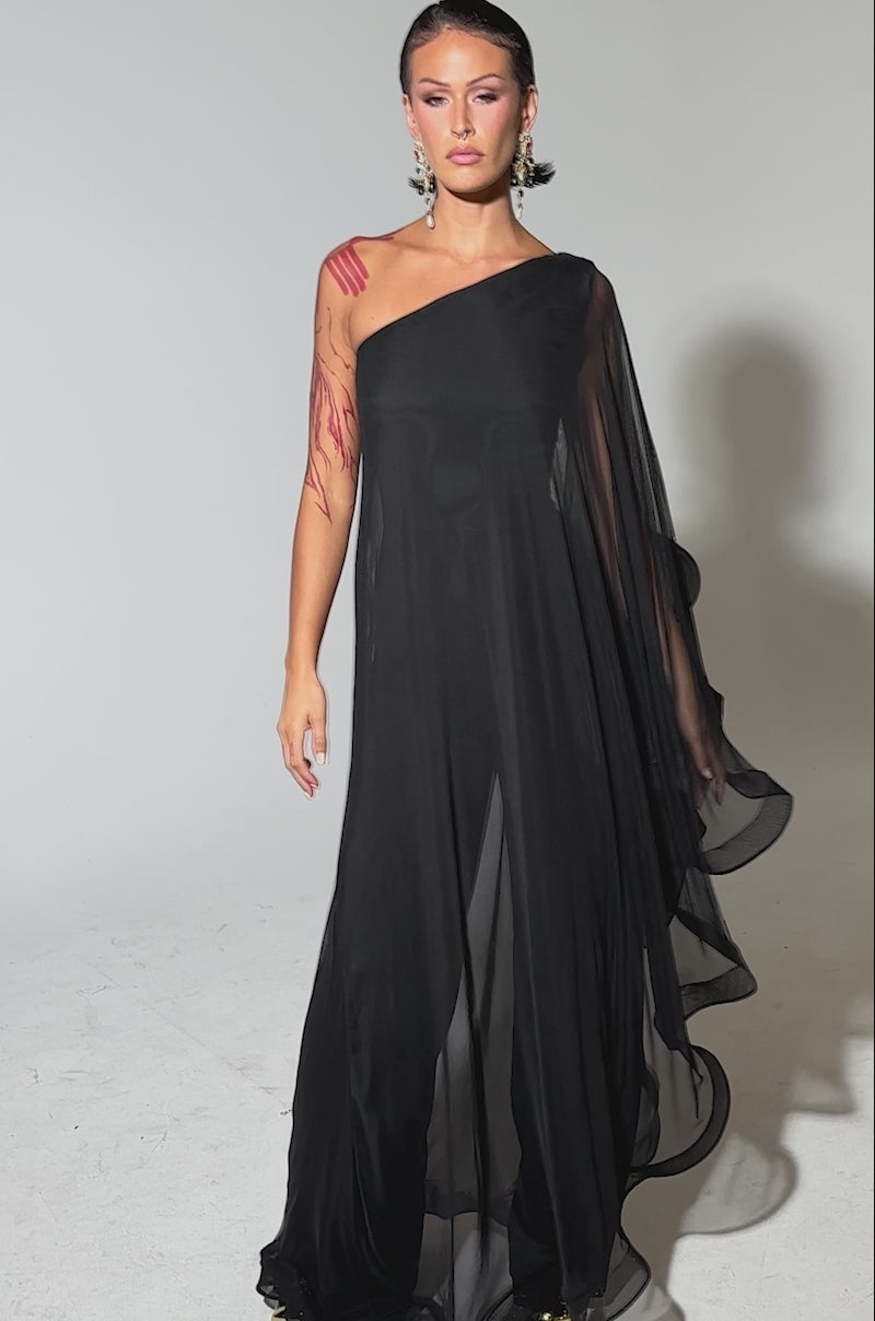 A video of a woman in the SANDO MESH JUMPSUIT IN BLACK, featuring sheer mesh fabric, ruffled details, and wide draping. She accessorizes with statement earrings and has slicked-back hair against a plain light-gray background.